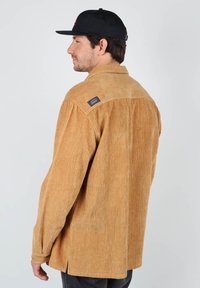 Chemise en velours côtelé de couleur marron clair, avec une finition texturée, des poignets boutonnés et une étiquette logo sur l'épaule. Stylée avec une casquette noire.