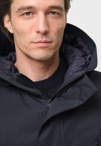 Svart vadderad jacka med hög krage, utrustad med dragkedja framtill och en ribbad stickad tröja under, slät tyg och en strukturerad huva.