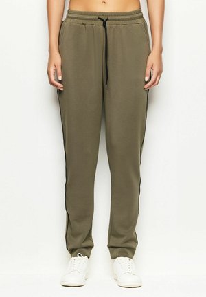 Persona che indossa pantaloni jogger verde oliva con strisce nere laterali e sneakers bianche, in piedi davanti a uno sfondo bianco.