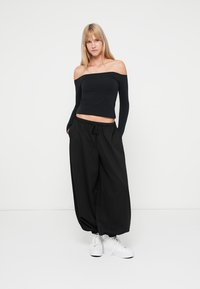 Mustmust kaelusega must topp ja laiad mustad püksid, mis on valmistatud kerge kangaga. Valged tossud. Ühtlane taust.