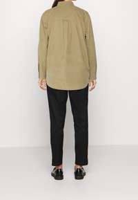 Camisa de pana beige con un pliegue en la parte trasera, mangas largas y un dobladillo redondeado, combinada con pantalones negros y zapatos negros brillantes.