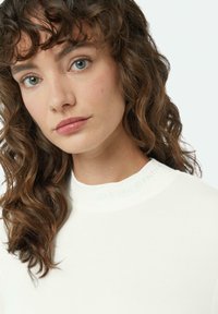 Biała bluzka z wysokim kołnierzem z subtelnym napisem "CASUAL IDENTITY" na kołnierzu. Gładka faktura i dopasowany krój podkreślają falowane, średniej długości włosy.
