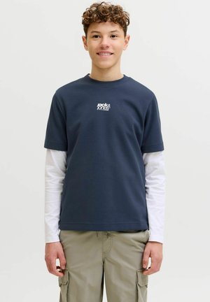 Młoda osoba o kręconych włosach, uśmiechająca się, w granatowej koszulce Jack & Jones na białej koszuli z długim rękawem oraz beżowych spodniach typu cargo.