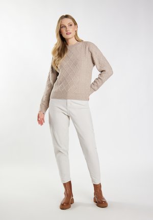 Vrouw staat met hand op heup, draagt een beige kabelgebreide trui, witte broek en bruine enkellaarzen tegen een effen achtergrond.