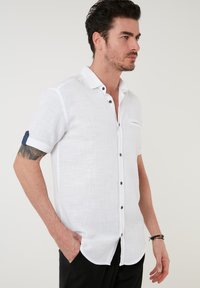 Camisa blanca de lino de manga corta con cuello abotonado, botones negros y un detalle de lunares azules en los puños enrollados. Tejido texturizado.