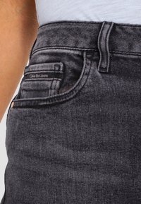 Pantalones vaqueros negros de mezclilla con cintura alta, que cuentan con un pequeño bolsillo delantero y una etiqueta de marca en la pretina. Textura suave y ajuste entallado.