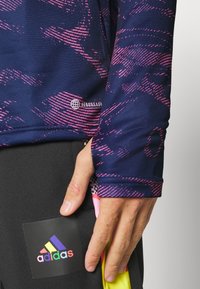 Laivastonsininen pitkähihaispaita, jossa on vaaleanpunaisia abstrakteja kuvioita ja Aeroready-brändäys. Lähikuva kädestä mustien housujen päällä, joissa on monivärinen Adidas-logo.