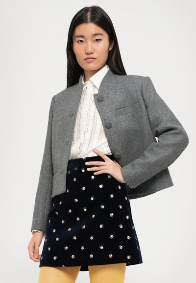 Veste cropped en mélange de laine grise avec des boutons ronds, associée à un chemisier blanc à bordure en dentelle, et une mini-jupe marine avec broderies florales.