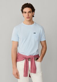 Jeune homme portant un t-shirt bleu clair avec un pull rose noué autour de la taille, debout devant un fond gris uni.