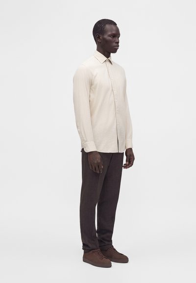 Homme debout avec une expression neutre portant une chemise crème à manches longues boutonnée, un pantalon foncé et des chaussures marron sur un fond clair uni.