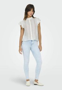 Blouse en dentelle blanche avec un col à volants haut et des manches courtes à volants, portée avec un jean skinny bleu clair et des chaussures slip-on crème.