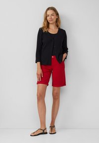 Vrouw staat tegen een effen grijze muur, gekleed in een zwarte knooptrui, een zwart topje, rode knielange shorts en zwarte platte sandalen.