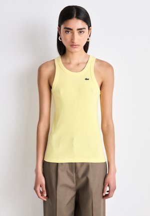 Débardeur jaune à côtes avec un col rond et un petit logo vert sur le côté gauche. Porté avec un pantalon plissé marron.