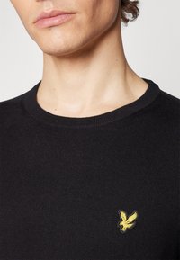 Pullover nero a maglia con scollatura a girocollo a coste e un piccolo emblema giallo a forma di aquila sul lato sinistro del petto. Texture liscia e design aderente.