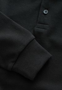 Schwarzes Poloshirt aus strukturiertem Stoff, mit gerippten Bündchen und einer Knopfleiste mit zwei Knöpfen sowie einer einzelnen Brusttasche.