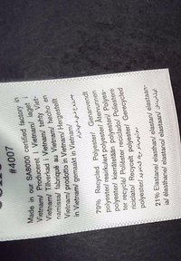 Odevná etiketa uvádzajúca výrobok vyrobený vo Vietname s obsahom látky: 79 % recyklovaný polyester a 21 % elastan, v niekoľkých jazykoch vrátane angličtiny a arabčiny.