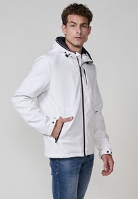 Jachetă albă din softshell, cu glugă, fermoar frontal și căptușeală contrastantă în nuanță de navy. Include buzunare frontale și un finisaj texturat al țesăturii.