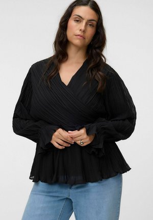 Femme aux longs cheveux foncés portant un chemisier noir plissé à manches longues et un jean bleu, debout les mains jointes à la taille.