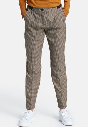 Personne portant un pantalon fuselé marron à carreaux, un haut à manches longues moutarde et des baskets blanches à lacets, debout les mains dans les poches.