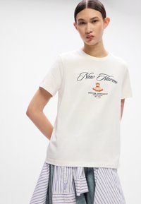 White cotton t-shirt with short sleeves, featuring "New Haven" in cursive and a graphic emblem. Worn over a blue and white striped shirt.