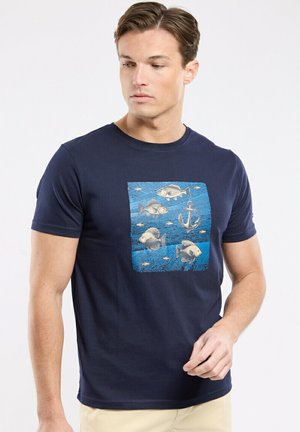 Homme portant un t-shirt bleu marine avec un graphique carré bleu représentant cinq poissons et une grande ancre sur la poitrine, regardant sur le côté.