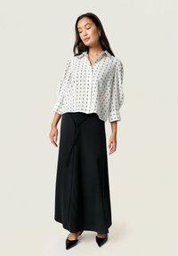 Blouse blanche à motifs avec des manches courtes bouffantes, associée à une longue jupe noire à taille nouée. Des chaussures noires à bout pointu complètent la tenue.