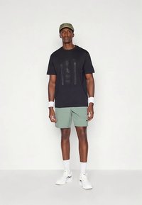 Μαύρο κοντομάνικο T-shirt με ανάγλυφο λογότυπο "BOSS", συνδυασμένο με ανοιχτοπράσινα σορτς. Αξεσουάρ περιλαμβάνουν πράσινο καπέλο και λευκές περιβραχιόνες.