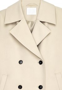 Beige double-breasted coat with wide lapels, four black buttons, two side pockets, and a visible NA-KD brand label inside the collar.