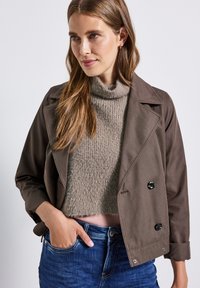 Veste marron croisée avec de grands boutons noirs, assortie à un col roulé en tricot gris clair. Le mannequin porte un jean bleu et pose avec une main dans sa poche.