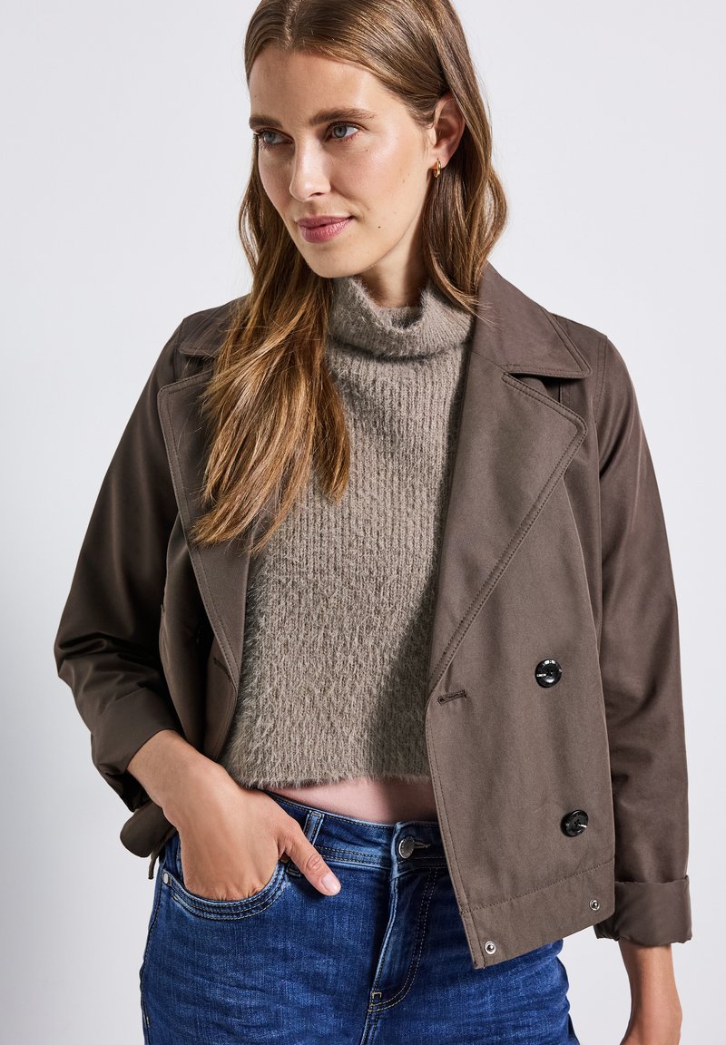 Veste marron croisée avec de grands boutons noirs, assortie à un col roulé en tricot gris clair. Le mannequin porte un jean bleu et pose avec une main dans sa poche.