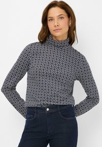 Maglione a collo alto blu navy in tessuto elastico con un motivo geometrico bianco. Maniche lunghe e silhouette aderente. Abbinato a jeans scuri.