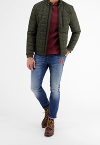 Vadderad grön jacka med hög krage över en vinröd stickad tröja, i kombination med blå jeans och bruna snörkängor.