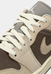 Καφέ και μπεζ αθλητικό παπούτσι με υφή δέρματος, κορδόνια από βαμβάκι και γκρι υφασμάτινες λεπτομέρειες. Διαθέτει ραμμένο σχέδιο και ορατό λογότυπο.