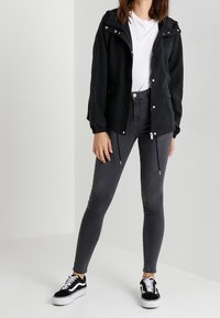 Giacca leggera nera con cappuccio, chiusura con zip e coulisse. Abbinata a jeans grigi slim-fit e sneaker nere con suole bianche.
