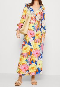 Vestido maxi floral en colores vibrantes, presenta un diseño envolvente, mangas largas y una cintura ceñida. Se acompaña de un bolso de paja trenzada y sandalias.