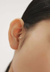Pequeño pendiente de botón con una gema redonda y clara engastada en una base metálica, llevado en el lóbulo de una oreja con piel suave y cabello lacio y oscuro.