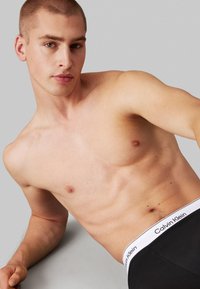 Sorte boxer briefs med en hvit Calvin Klein-logo på midjebåndet, vist på en modell med barbert hode og en nøytral grå bakgrunn.