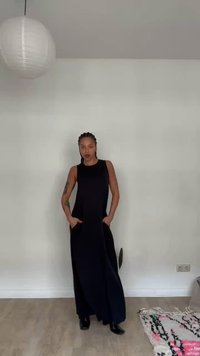 Γυναίκα με πλεξούδες που φοράει μακρύ μαύρο αμάνικο φόρεμα στέκεται σε ξύλινο πάτωμα μέσα σε εσωτερικό χώρο δίπλα σε μοτίβο χαλί και κρεμαστό φωτιστικό από χαρτί.