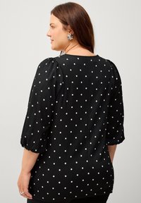 Zwarte blouse met witte stippen, gestructureerde stof, pofmouwen en een ronde halslijn, gezien vanuit de achterkant.