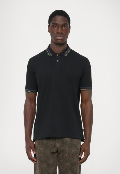 Hombre con una camiseta polo negra con rayas multicolores en el cuello y las mangas, combinado con pantalones marrones de estampado floral, de pie frente a un fondo blanco.
