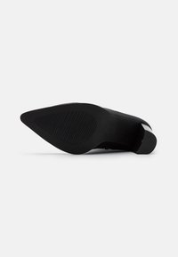 Zapato de tacón alto negro con punta afilada y suela de goma texturizada, que presenta un empeine de cuero suave y un diseño minimalista.