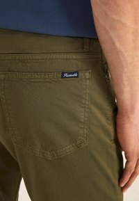 Pantalones verde oliva con una textura suave, que cuentan con un bolsillo trasero y una etiqueta azul con texto blanco. El diseño incluye detalles de costura simple.