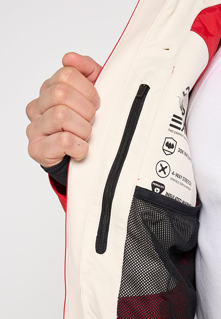 Eine Hand hält eine rot-weiße Jacke geöffnet, die eine aufgeschlossene Innentasche mit Reißverschluss und eine schwarze Netztasche mit Pflege-Symbolen zeigt.