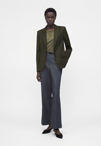 Femme portant un blazer olive, un top vert assorti, un pantalon large gris taille haute et des ballerines noires pointues, debout devant un fond uni.