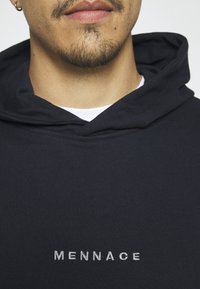 Černá mikina s kapucí s texturovaným povrchem, vybavená přední kapsou a vyšitým logem "MENNACE" v bílé barvě. Součástí je kapuce se stahovací šňůrkou.