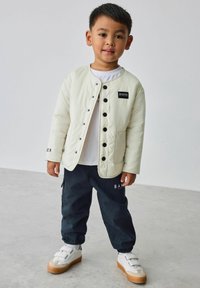 Hellcremefarbene gesteppte Jacke mit schwarzen Knöpfen und Logo, kombiniert mit dunkelblauen Hosen mit Logo und weißen Sneakern mit Klettverschlüssen.