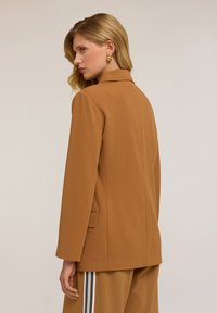 Blazer marrone sagomato con un design strutturato, maniche lunghe e uno spacco posteriore singolo. Il tessuto appare liscio con una leggera texture.