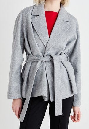 Femme portant un manteau en laine gris clair ceinturé, par-dessus un haut rouge et un pantalon noir, sur un fond blanc uni.