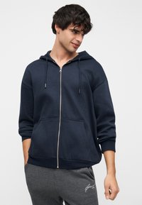 Φούτερ με φερμουάρ σε ναυτικό μπλε, φτιαγμένο από μαλακό ύφασμα, με κουκούλα ρυθμιζόμενη με κορδόνι, μπροστινές τσέπες και ριμπ μανσέτες. Φοράται με γκρι φόρμα.