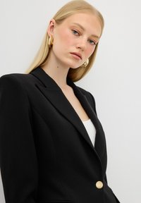 Blazer nero con un design strutturato, bottone dorato singolo e revers affilati. Il modello indossa una maglietta bianca, con lunghi capelli biondi e orecchini a cerchio dorati.
